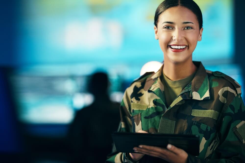 A person in a military uniform stands indoors with a smile, holding a tablet. The background is blurred but shows another veteran working at a desk with multiple screens. The atmosphere suggests a technology or command center.